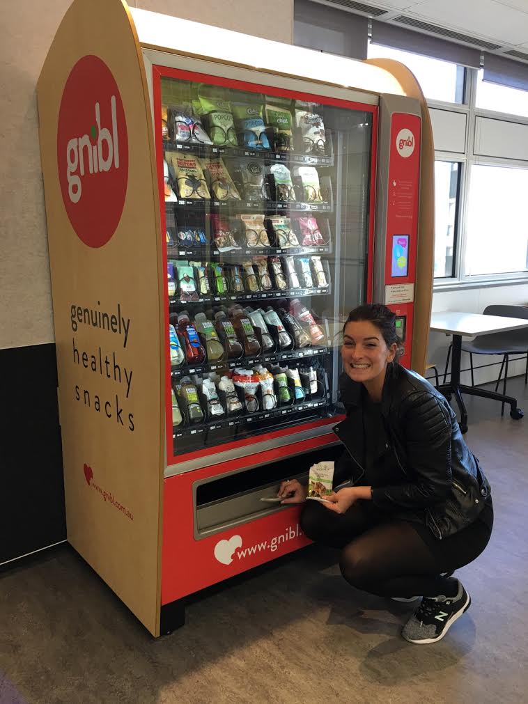 A happy customer crouched in front of a Gnibl combination vending machine after making a snack purchase.
