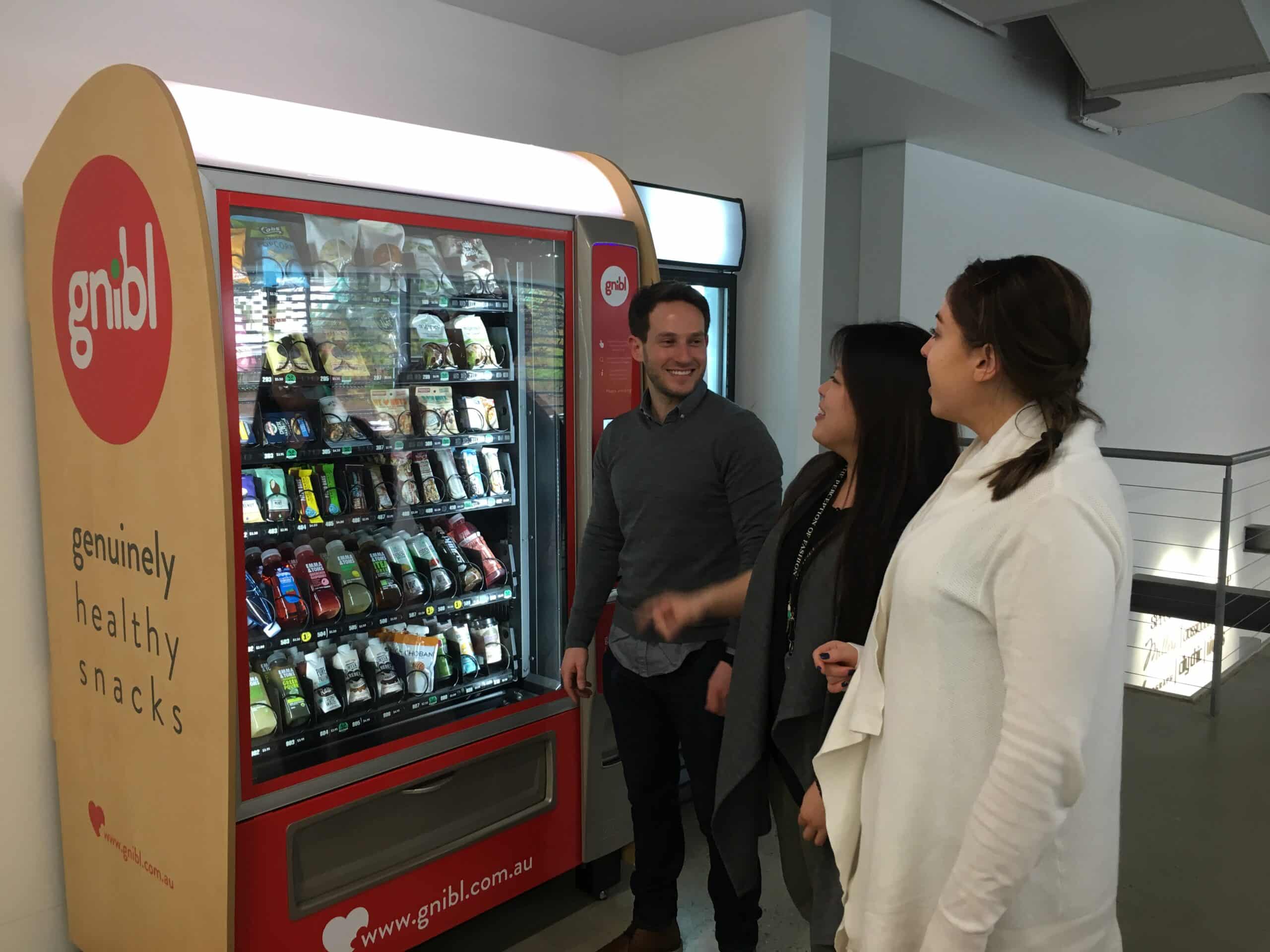 Gnibl Founder Nick Volpe with two women in front of a Gnibl vending machine in a workplace setting.