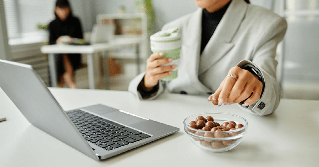 A worker snacking while holding a coffee cup and sitting in front of a laptop.